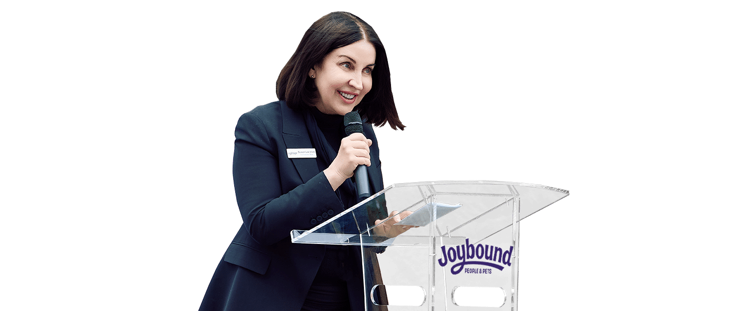 A woman speaking at a clear podium with a Joybound sign, wearing a dark blazer against a white background.